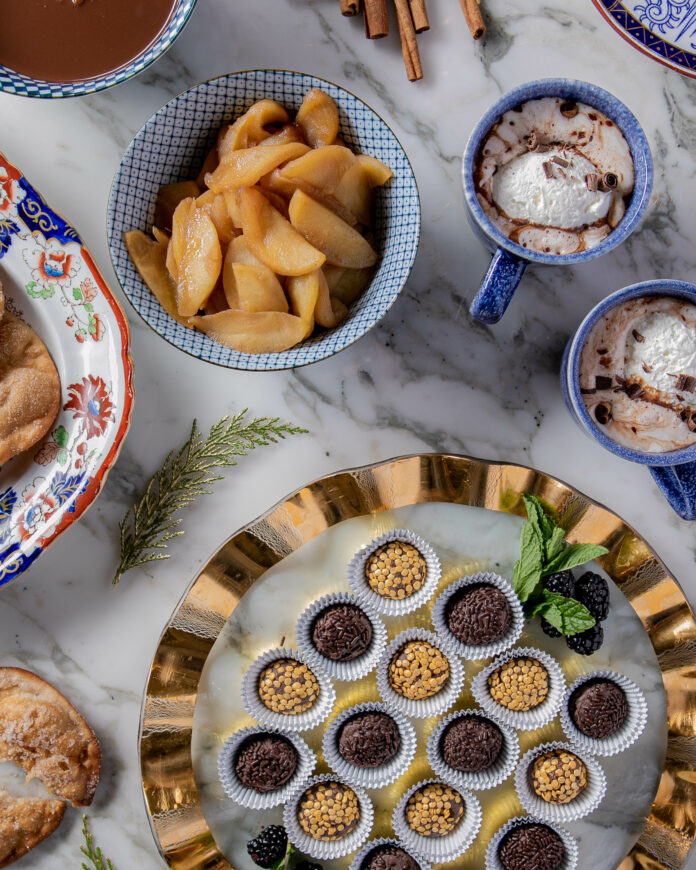 A variety of small snacks in a surrounding circle with small mugs and plates present. Brigadeiros