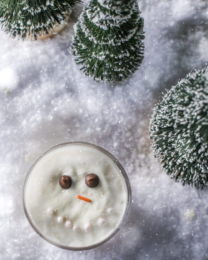 A Melted Snowman cocktail with an edible face sits in the middle of snow and trees.