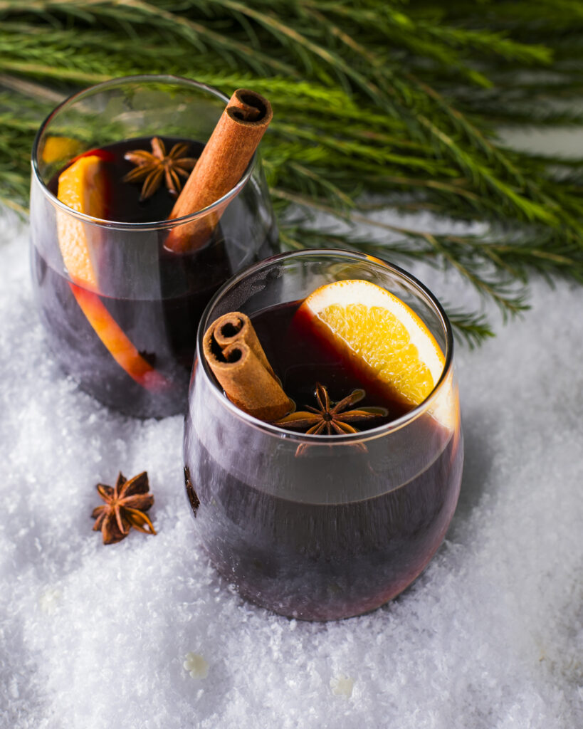 Two stemless wine glasses with mulled wine sitting on a snowy surface with pine greens, garnished with a cinnamon stick, star anise, and an orange slice.