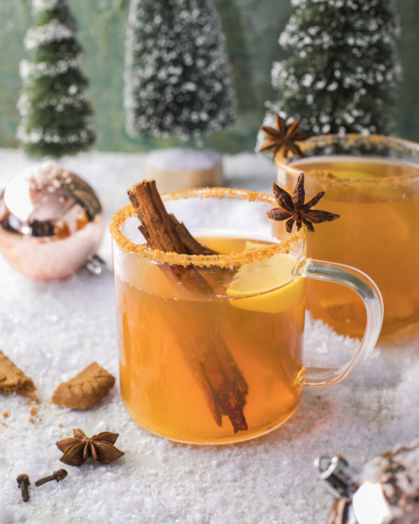 Two clear glass mugs with a ginger snap colored beverage garnished with a cinnamon stick, star anise, and lemon on a snowy surface with a shiny Christmas bulb and little pine trees in the background.