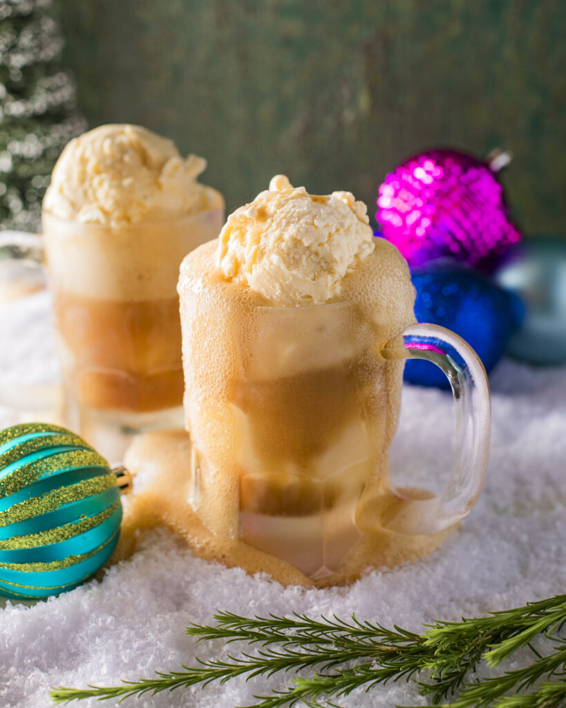 Two boozy root beer floats I frosted mugs overflowing with frothy root beer on a snowy surface with colorful Christmas ornaments and greenery.