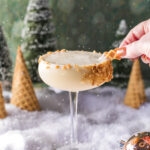 A creamy looking cocktail in a coupe glass with a crushed waffle cone rim on a snowy surface with cone trees and pine trees in the background.