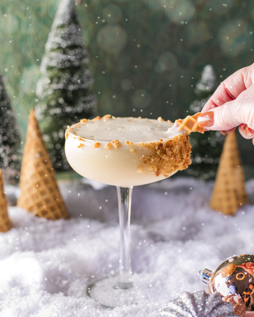 A creamy looking cocktail in a coupe glass with a crushed waffle cone rim on a snowy surface with cone trees and pine trees in the background.