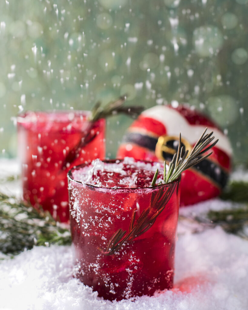 Two red rocks glasses with a spring of rosemary garnish, sitting on a snowy surface with a round Christmas ball that. looks like Santa's outfit.