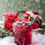 Two red rocks glasses with a spring of rosemary garnish, sitting on a snowy surface with a round Christmas ball that. looks like Santa's outfit.