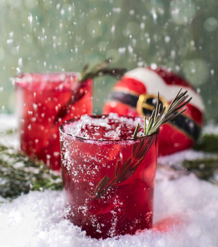 Red Ryder Punch Two red rocks glasses with a spring of rosemary garnish, sitting on a snowy surface with a round Christmas ball that. looks like Santa's outfit.