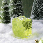 A green cocktail in a clear textured glass with dill "whisker "garnish to represent the Grinches hair, sitting on a pile of snow with a green shiny Christmas bulb and pine trees in the background, appropriately titled Mean and Green.