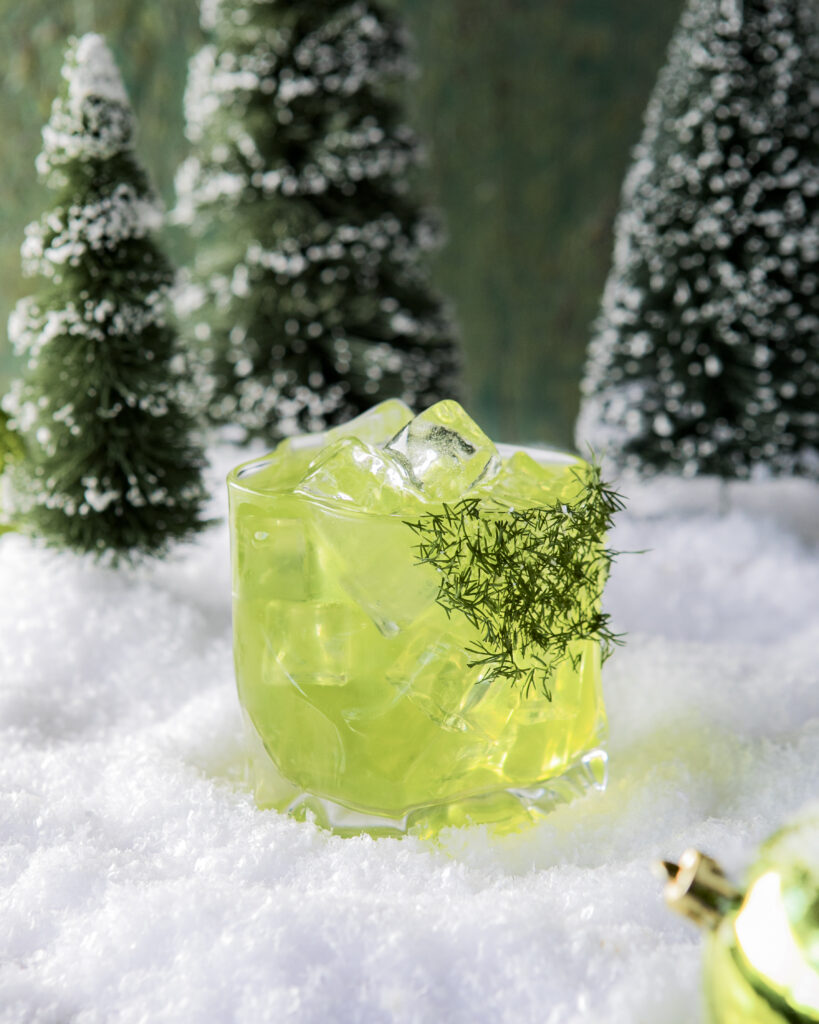A green cocktail in a clear textured glass with dill "whisker "garnish to represent the Grinches hair, sitting on a pile of snow with a green shiny Christmas bulb and pine trees in the background, appropriately titled Mean and Green.