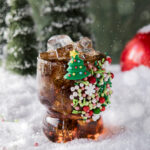 A snowy surface with a cola cocktail in a clear glass adorned with lots of red, green, and white holiday candy sprinkles as garnish on the glass made to remind you of Buddy's breakfast in the move Elf.