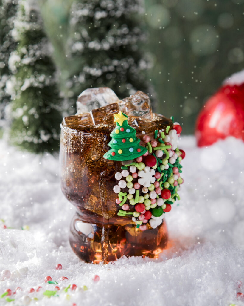 A snowy surface with a cola cocktail in a clear glass adorned with lots of red, green, and white holiday candy sprinkles as garnish on the glass made to remind you of Buddy's breakfast in the move Elf.