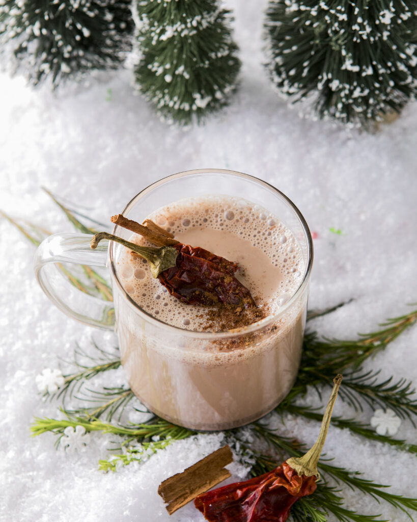 A clear mug of hot chocolate on a snowy surface with evergreen clippings and dried ancho chili peppers.