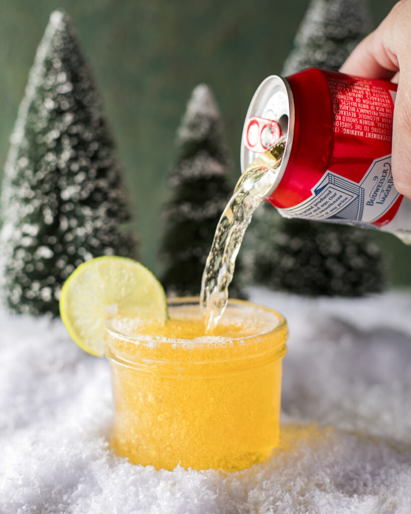 A red can of beer pours into a glass of Cousin Eddie's Trailer Fuel surrounded by snow and trees and garnished with a lime wheel.