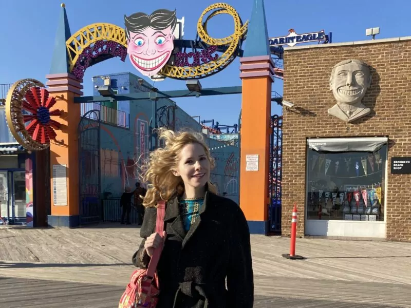 A woman with blonde hair stands in a jacket with a red bag in front of an amusement park fence.
