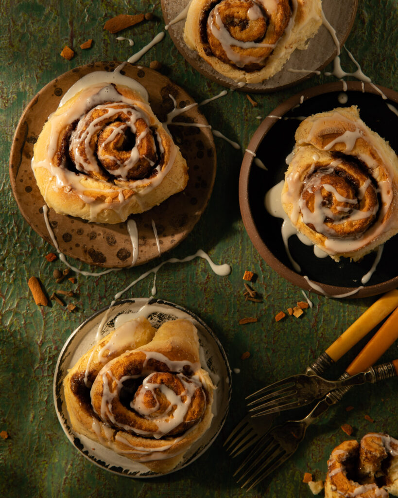 Five cinnamon rolls on five different small plates, drizzled with icing and garnished with broken cinnamon sticks on a green surface.