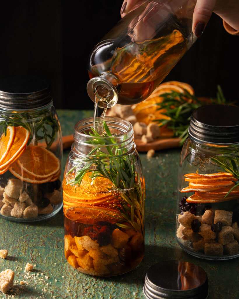 A mason jar filled with whiskey, oranges, sprigs of spices, and much more.