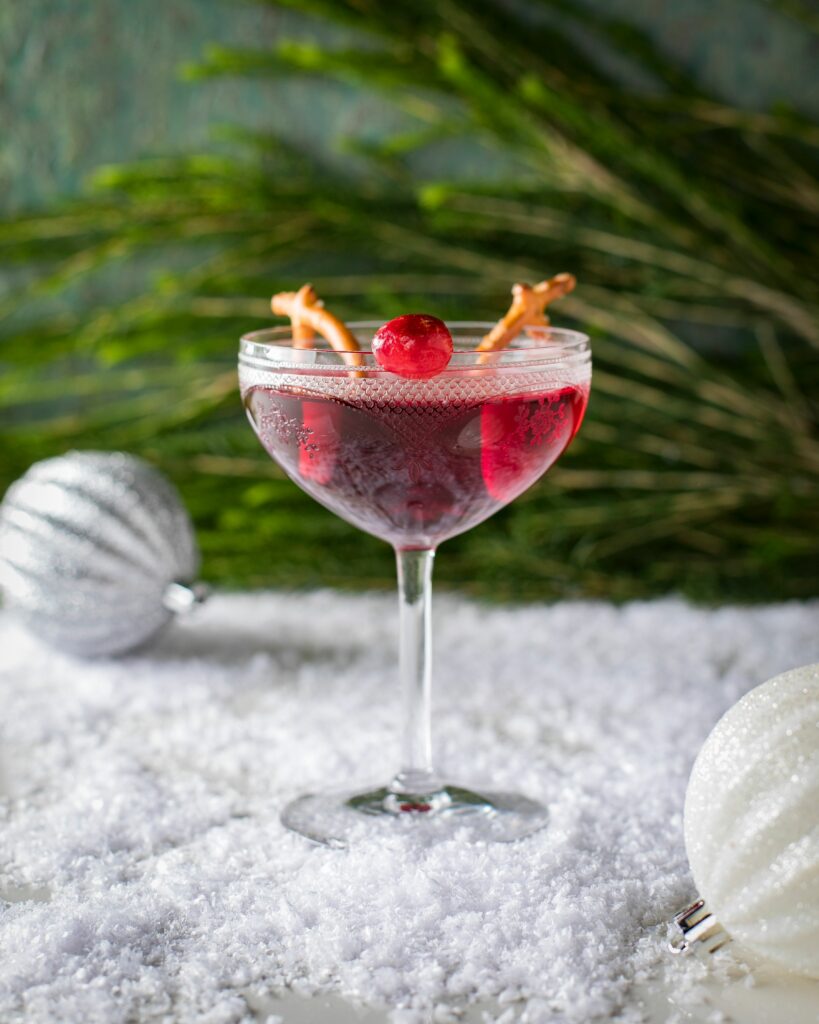 A cocktail glass filled with Rudolph's Red-Nose Punch topped with a cranberry and pretzels on the glass.