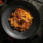 A spicy chicken sauce pasta dish homemade by Victoria Sande in a dark bowl surrounded by ingredients.