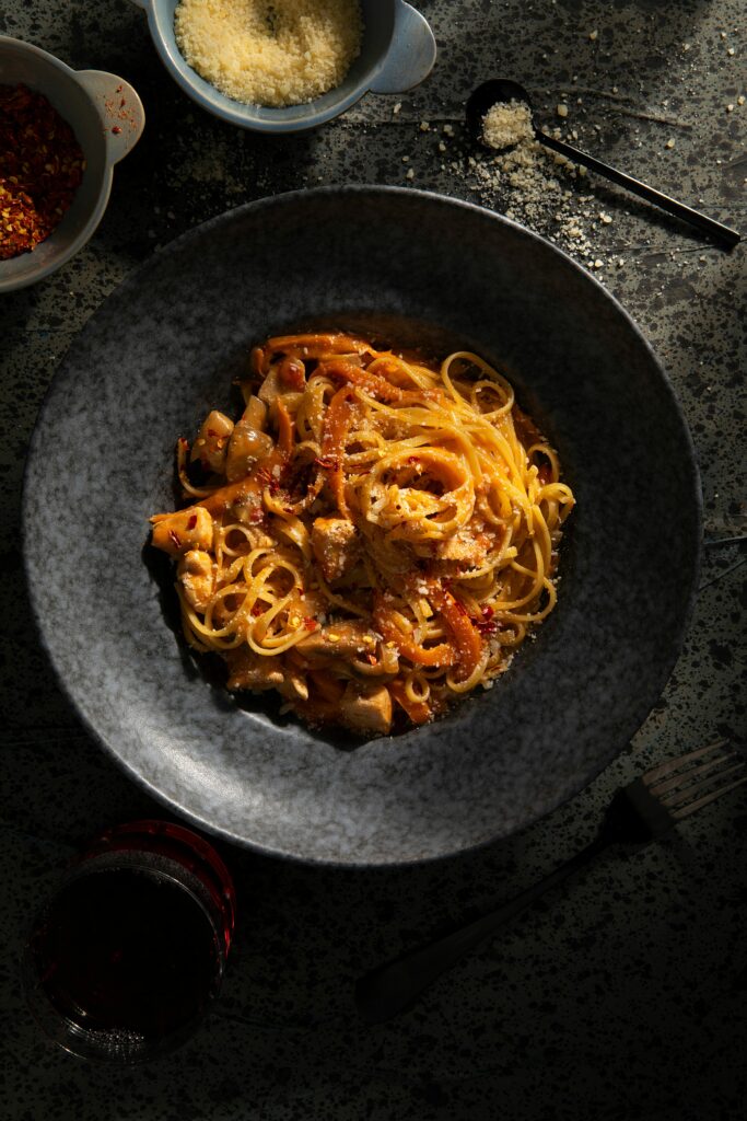 A spicy chicken sauce pasta dish Italian Homemade Recipe by Victoria Sande in a dark bowl surrounded by ingredients.