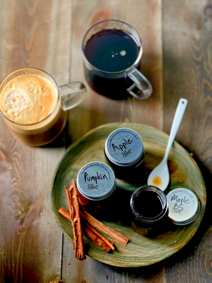 Three small mason jars filled with coffee syrups sit on a plate in front of two cups of coffee.