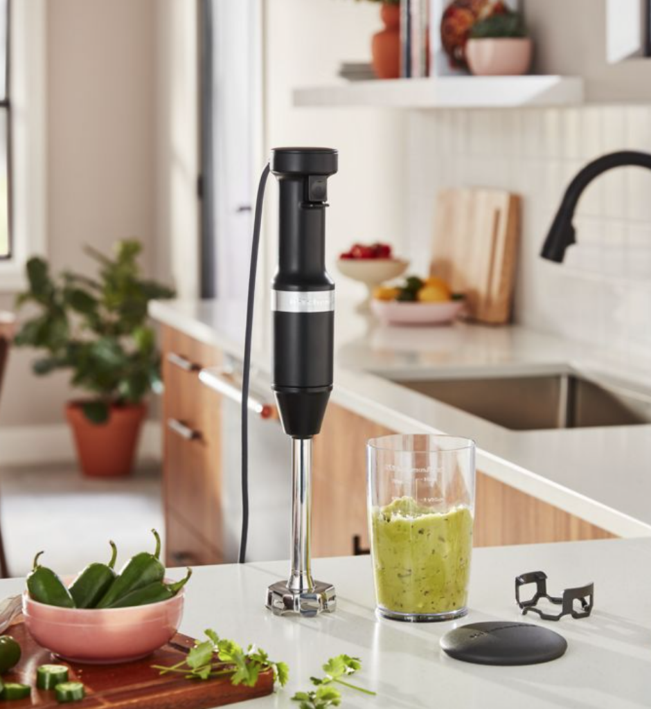 A black immersion blender sits on a marle countertop next to a mixed glass of chiles and a bowl of unblended chiles.