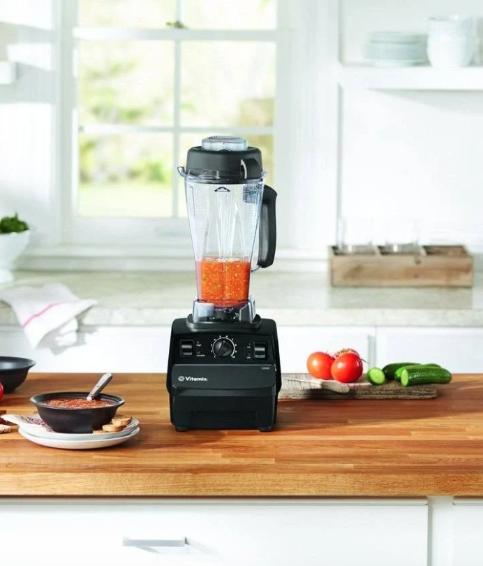 A Vitamix blender on a wooden counter with tomatoes, cumbers, and a bowl of soup also on the counter.