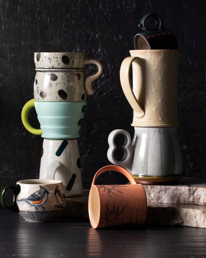 Two stacks of handmade mugs from an artist shop in Lawrenceville sit on a black background with a brown mug laying on its side in front of the stacks.