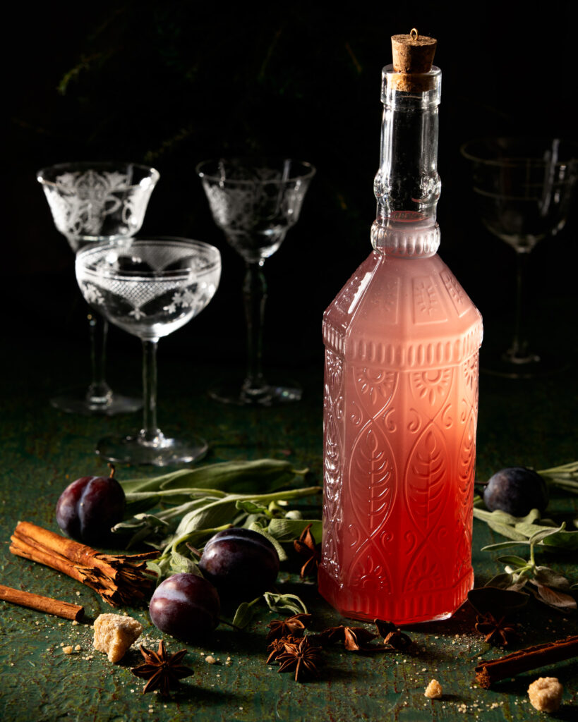 A bottle of Sugar Plum and Sage Infused Vodka sits on a table in front of three tall glasses.