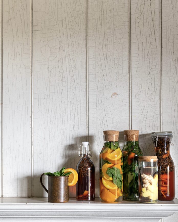 Five clear bottles of different sizes with infused liquors, sitting on the right of a shelf with a copper mug to the left.