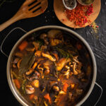 A stock pot with a variety of mushrooms and vegetables in a brown stock, with a wooden spoon in the upper left corner and some pepper and seasonings on a plate in the upper right corner.