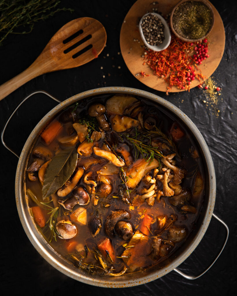 A stock pot with a variety of mushrooms and vegetables in a brown stock, with a wooden spoon in the upper left corner and some pepper and seasonings on a plate in the upper right corner.