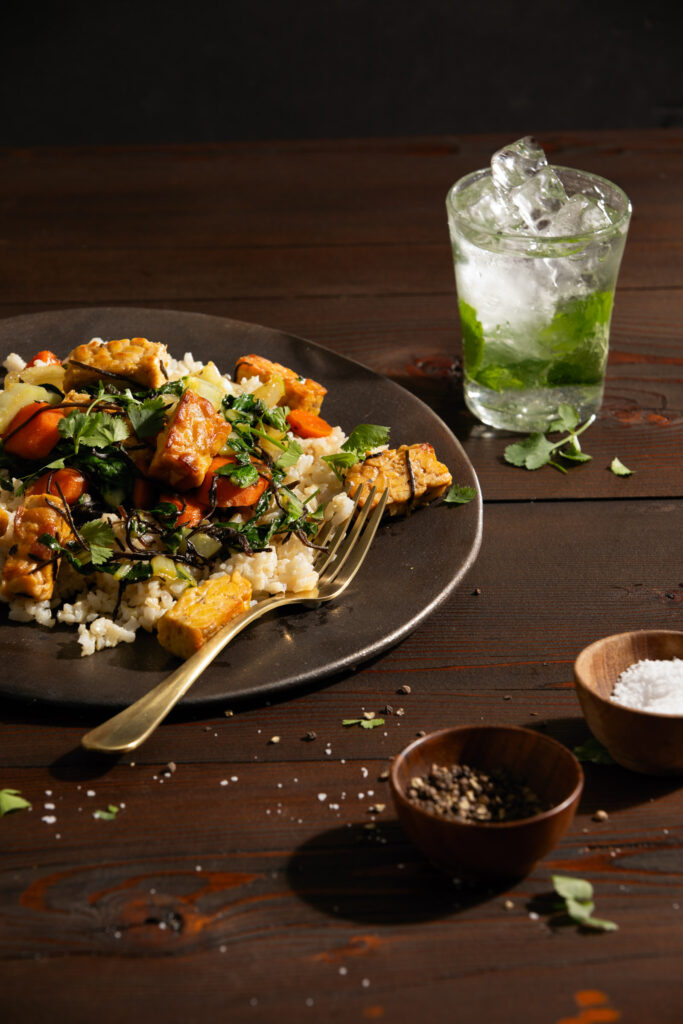 A grey plate sits off to the left side of the frame with a grains and greens, a gold fork sits on the right side of the plate. To the right of the frame is a green beverage in a glass. Tempeh & Bok Choy Stir Fry recipe for meatless monday.