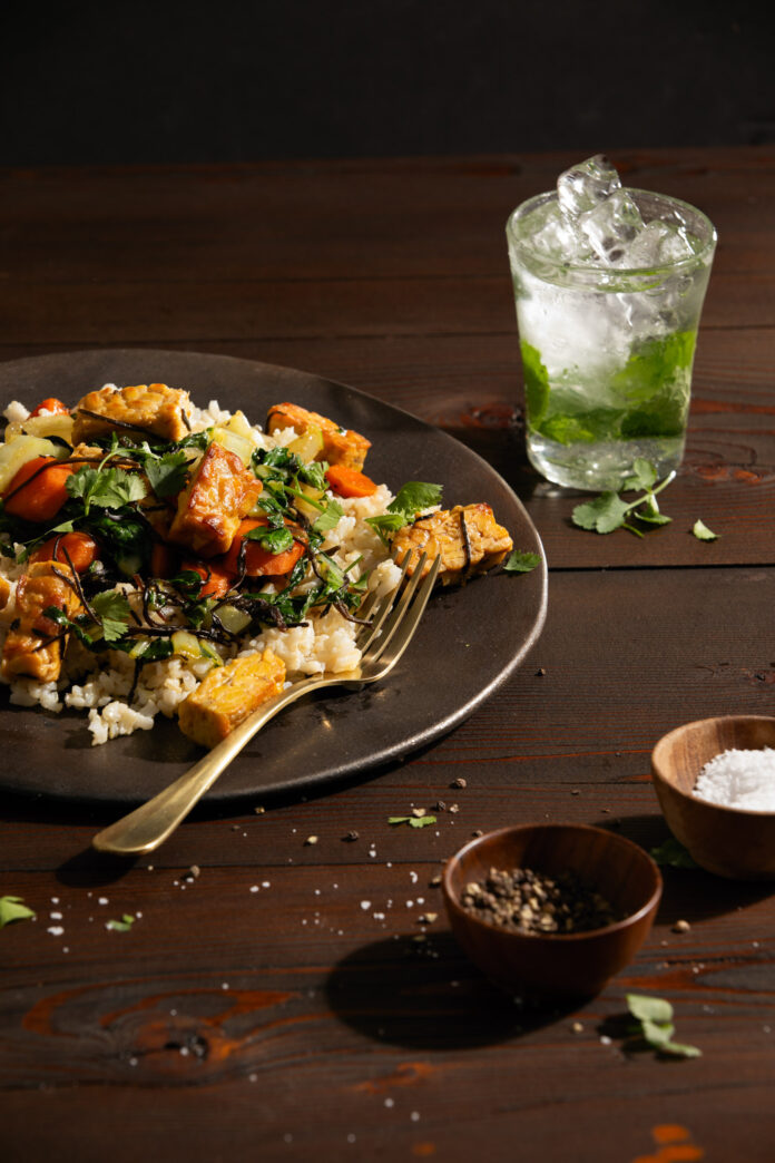 A grey plate sits off to the left side of the frame with a grains and greens, a gold fork sits on the right side of the plate. To the right of the frame is a green beverage in a glass. Tempeh & Bok Choy Stir Fry recipe