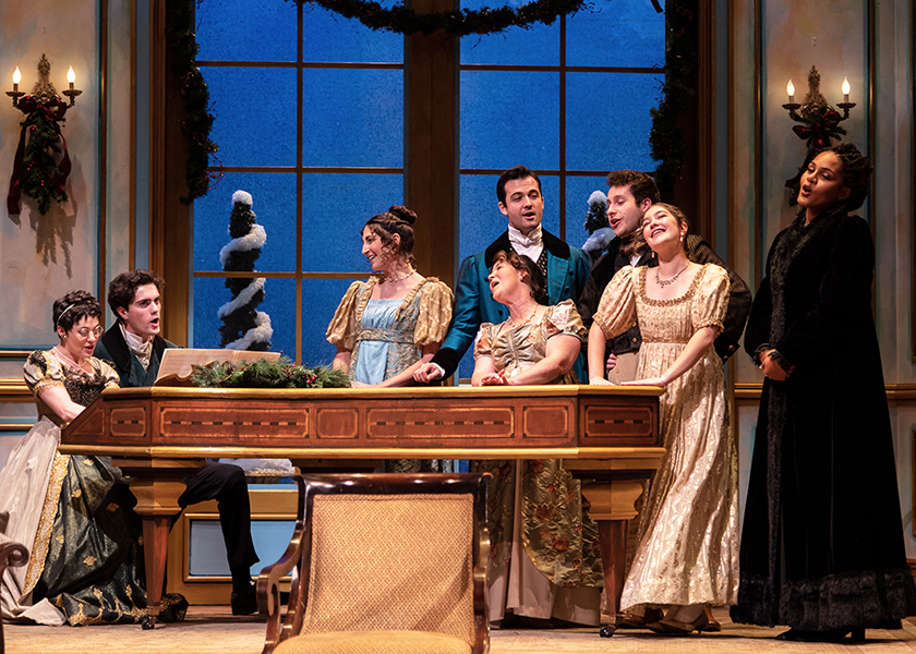 A ground of victorian characters gather around a piano to sing a song.