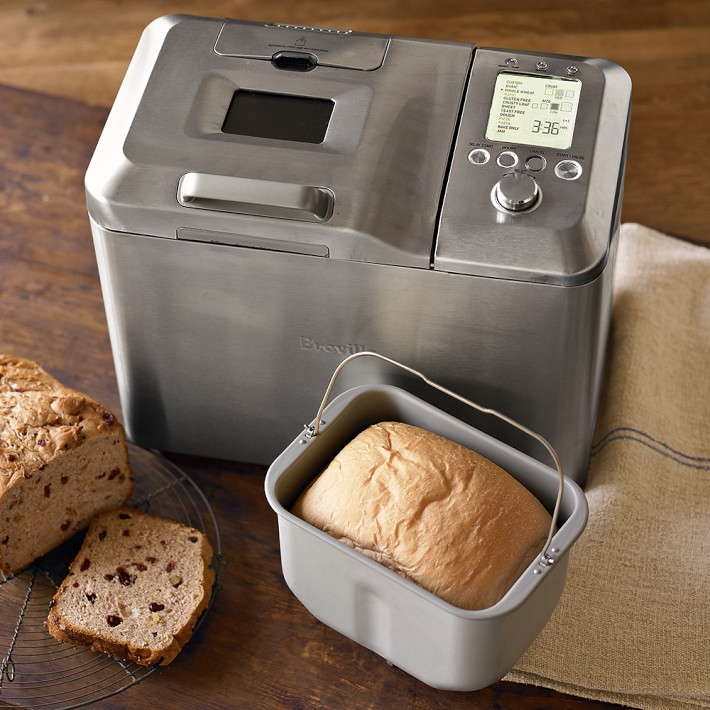 A silver bread maker with a loaf of bread inside on a table with more bread.