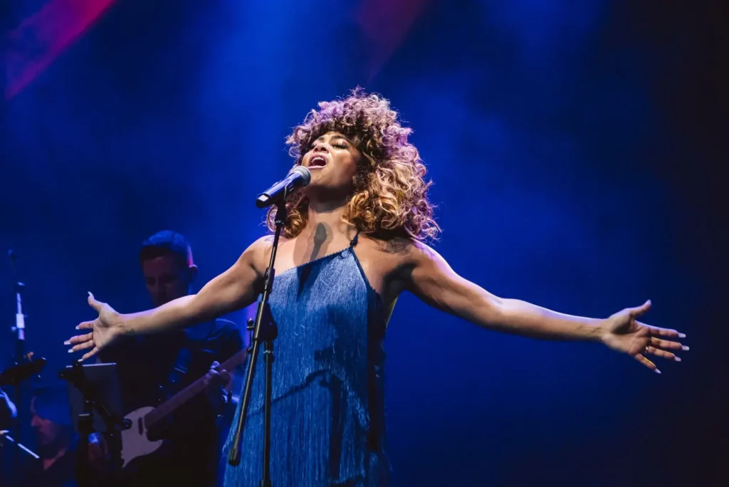 A woman portraying Tina Turner sings on stage in a blue dress to highlight the Pittsburgh production.