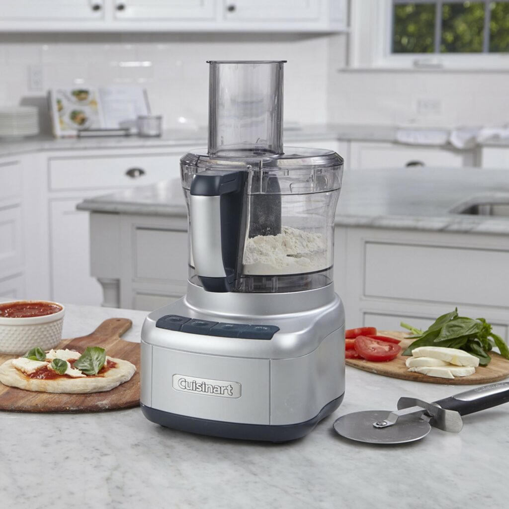 A silver food processor sits on a white marble table surrounded by fresh ingredients and a pizza cutter.
