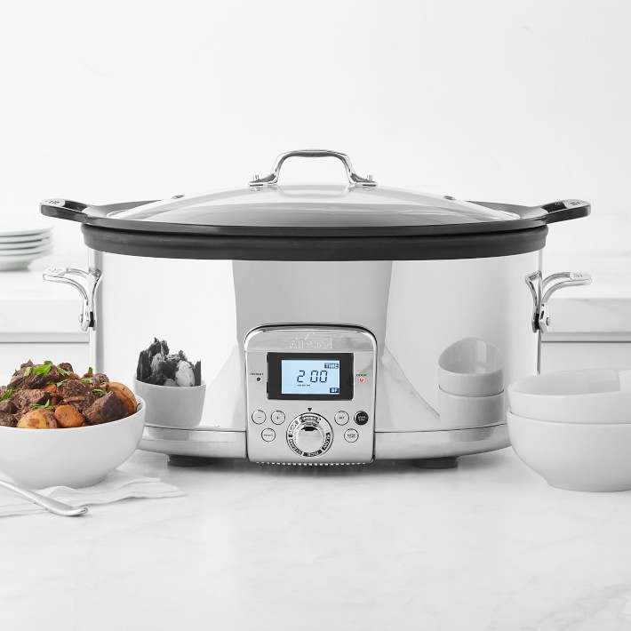 A silver slow cooker on a white table with a bowl of pot roast beside it and plates in the background.