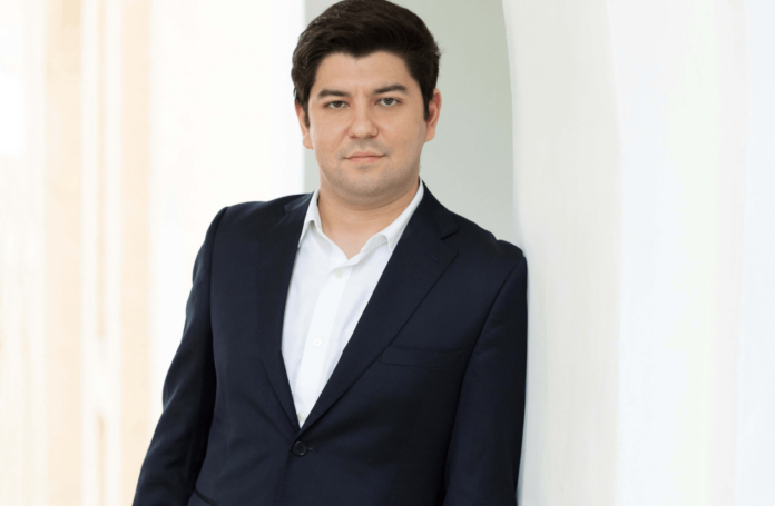 A man stands against a white pillar in a black suit jacket and white shirt from the Pittsburgh event guide.