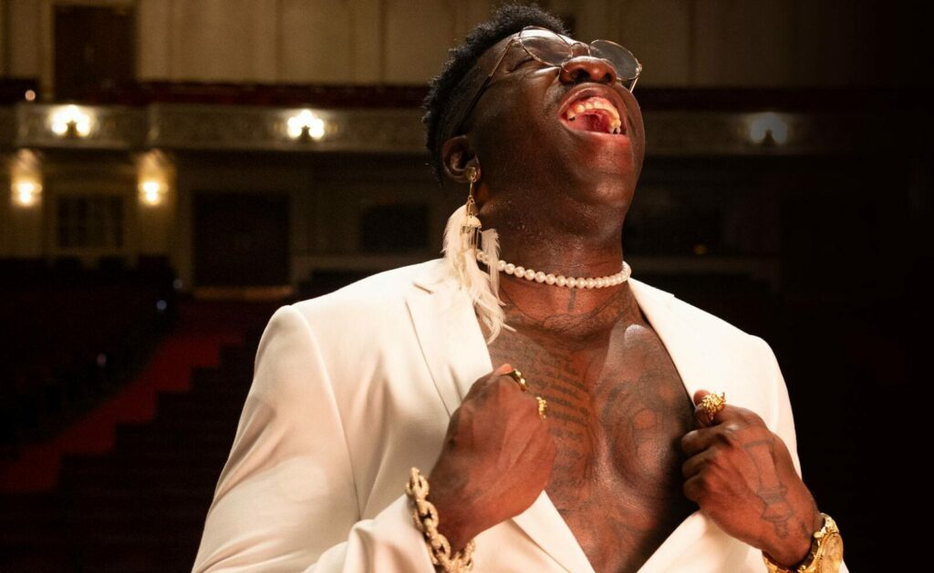 Rapper Frzy stands in a theater with his white shirt open and pearls around his neck.