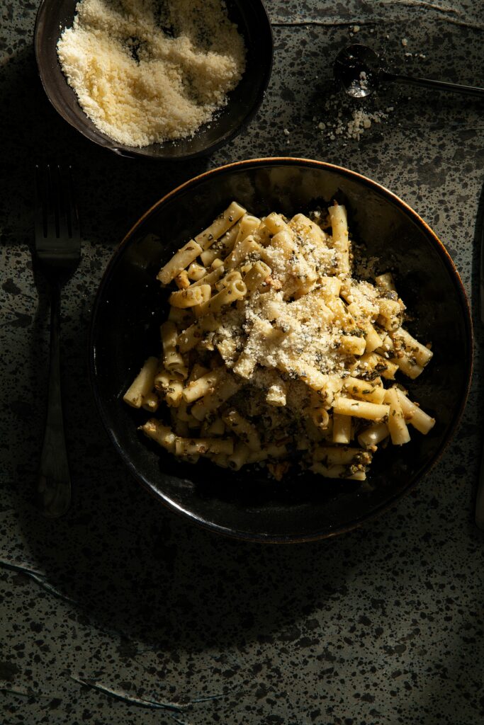 A dish of creamy pesto sauce pasta in a black bowl beside parmesan cheese made by Victoria Sande.