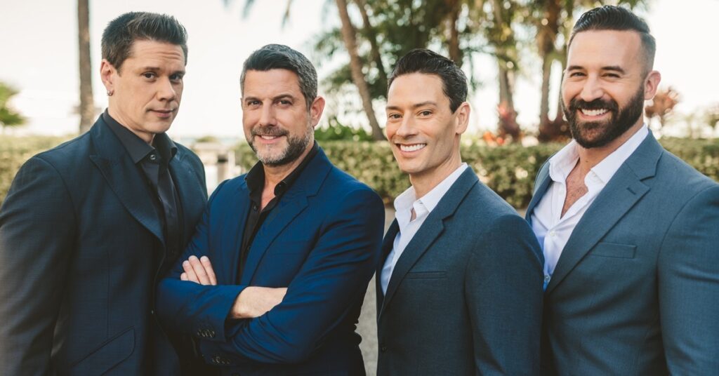 Four men in suits stand outside smiling at the camera. One has his arms crossed over his chest.