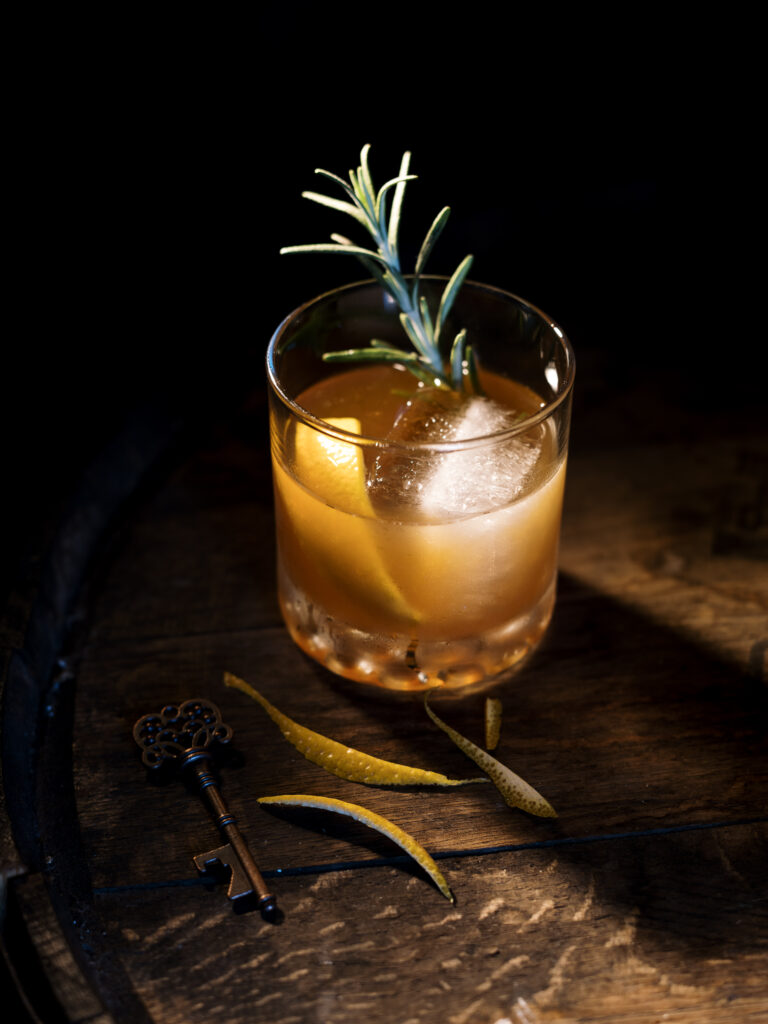 An amber drink in a short drinking glass with a large ice cube, orange peel, and sprig of rosemary.
