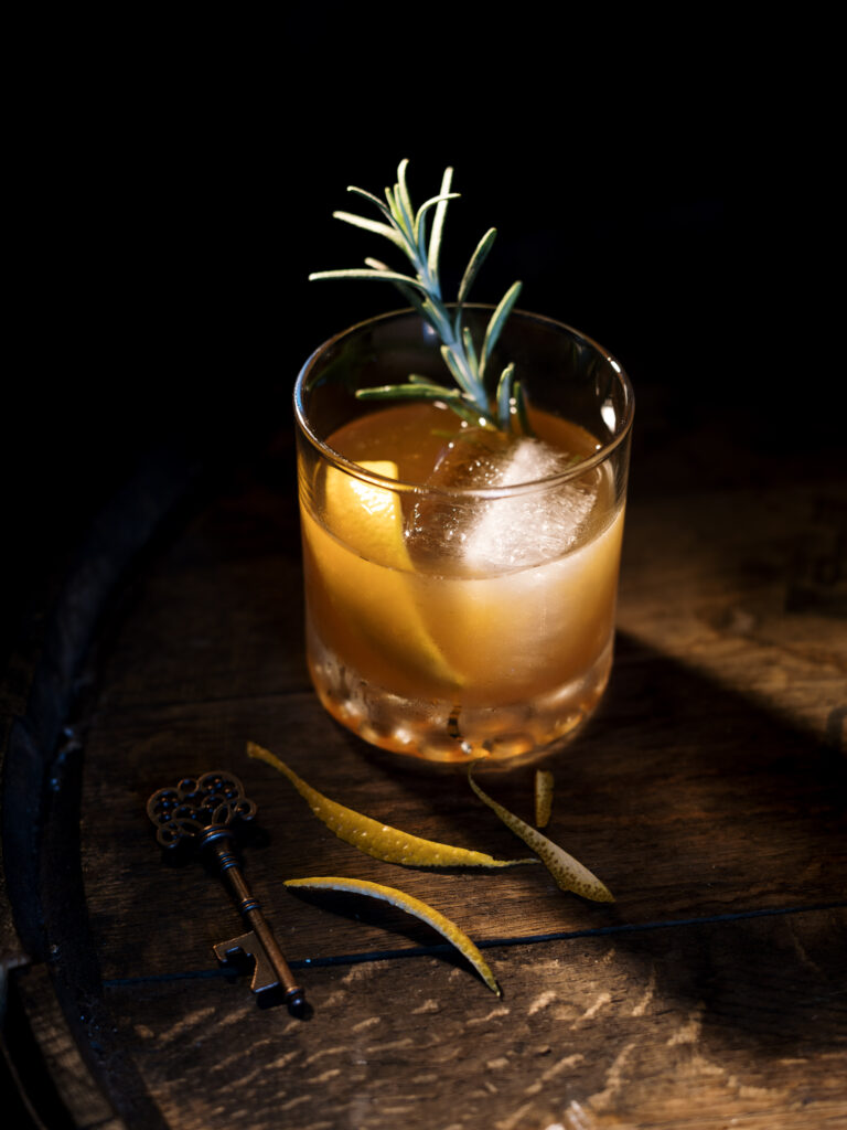 An amber drink in a short drinking glass with a large ice cube, orange peel, and sprig of rosemary.