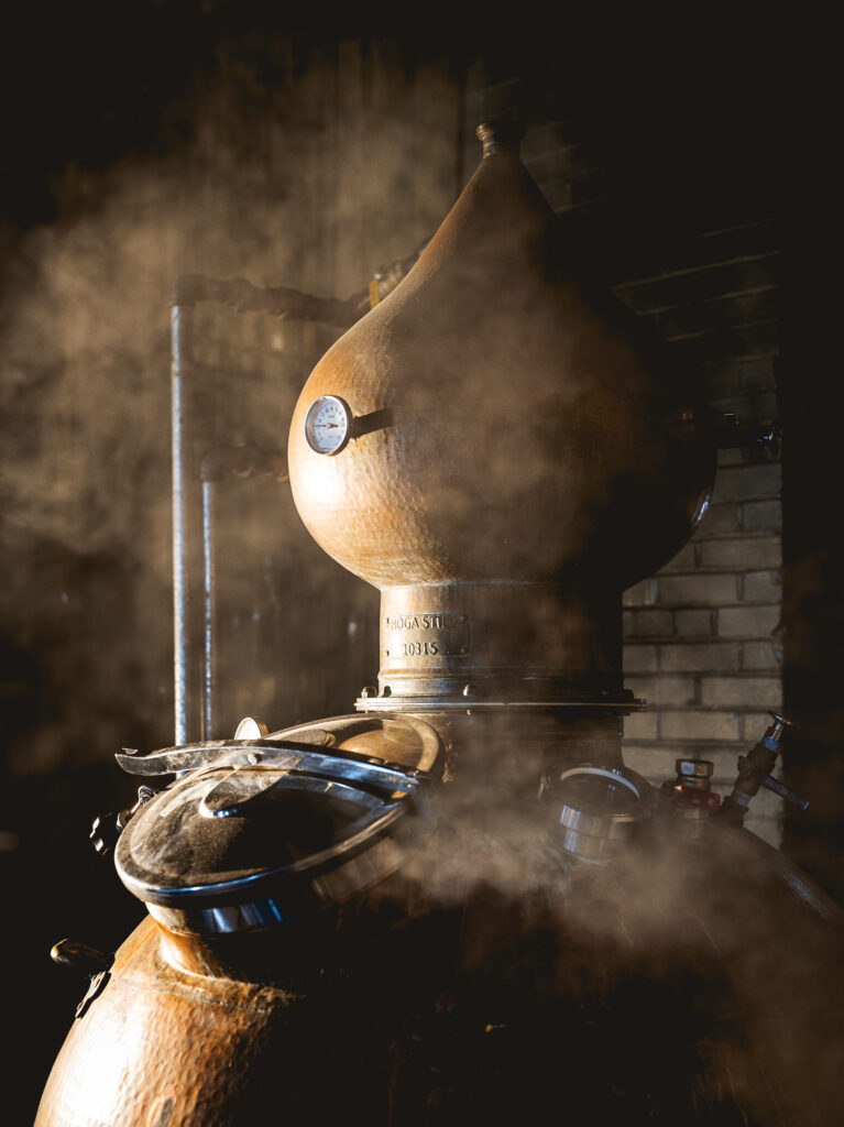 A distilling machine at a local Pittsburgh distillery with steam coming out all around it against a brick wall.