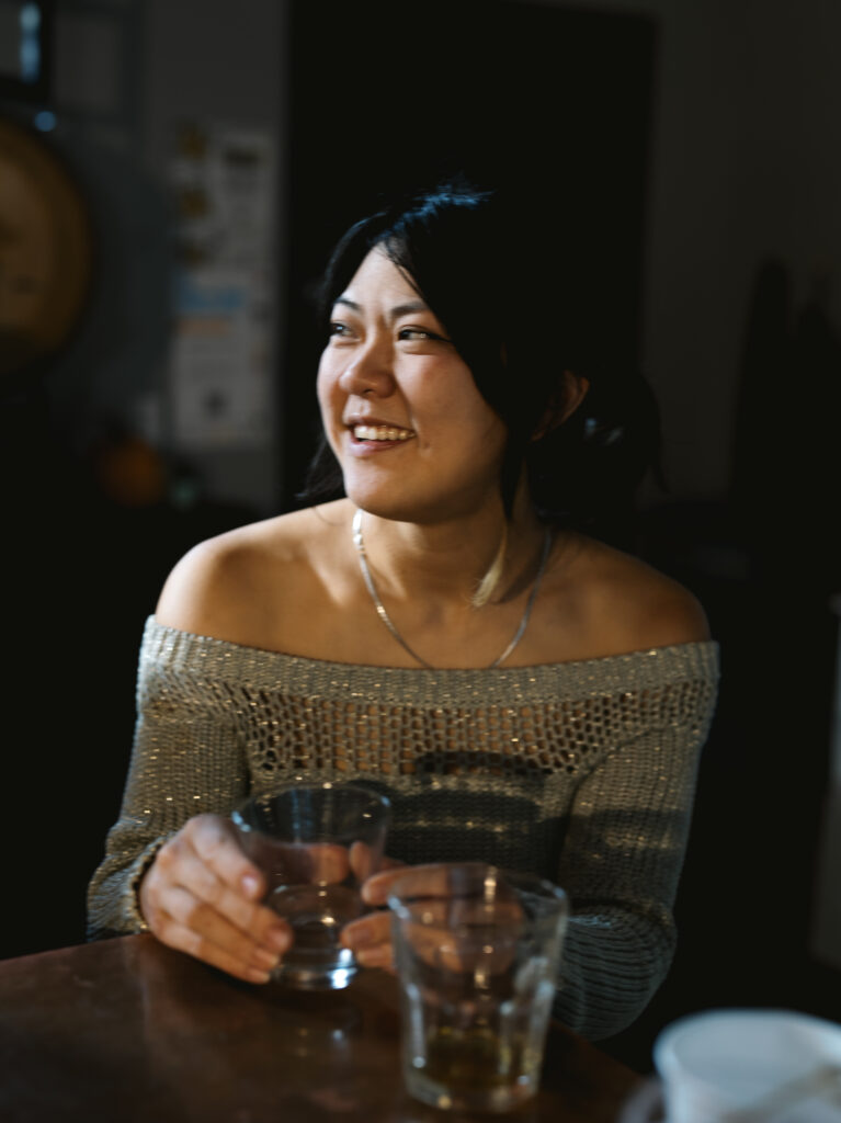 A woman with dark hair and a sweater off the shoulders sits at a table in a local Pittsburgh distillery with a glass in her hands.