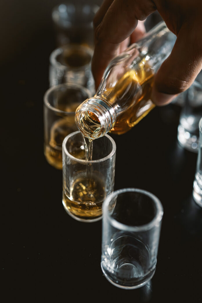 ©JOEYKENNEDY WKF 2-0005 Someone pours four shots of amber liquid on a black bar top.