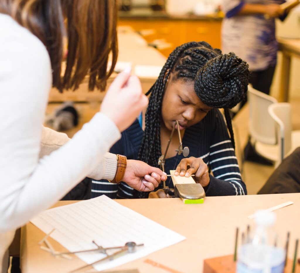 A woman helps a girl sitting at a wooden table working on a craft.