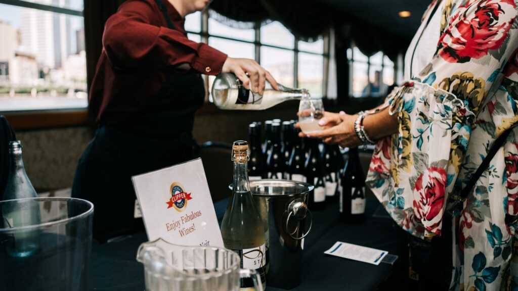 A person in a red jacket pouring another a glass of wine at the wine table aboard the Pittsburgh Gateway Clipper.