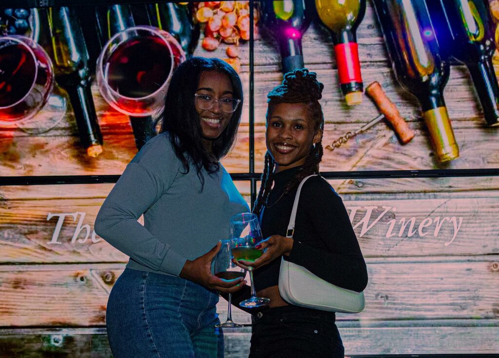 Two women stand in front of pictures of bottles of wines, holding wine in their own hands.