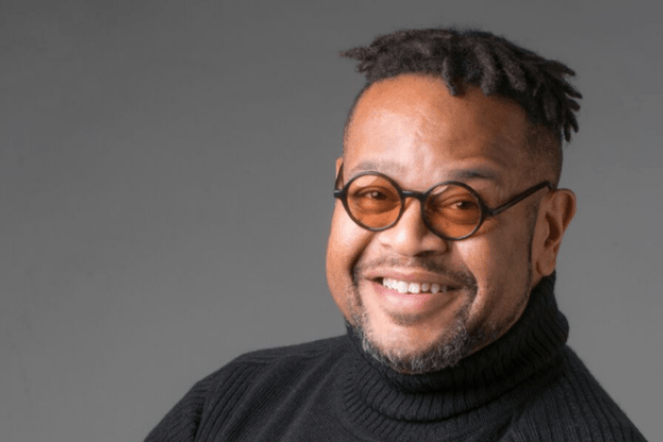 A man in small orange glasses and a black shirt smiles in front of a grey background.